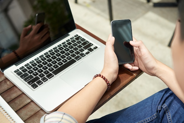 Unrecognizable man sitting with laptop in cafe and using smartphone