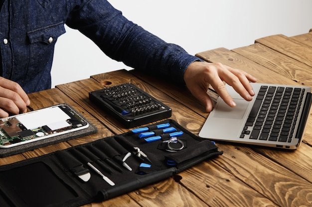 До неузнаваемости мужчина использует ноутбук, чтобы найти руководства по ремонту электронного устройства Сумка для инструментов и сломанный гаджет рядом с винтажным деревянным столом