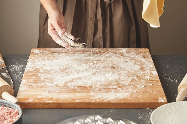 Бесплатное фото До неузнаваемости женщина насыпает белую муку на деревянную доску, держа на воздухе раскладывающее тесто для макарон или пельменей. пошаговая инструкция по приготовлению равиоли
