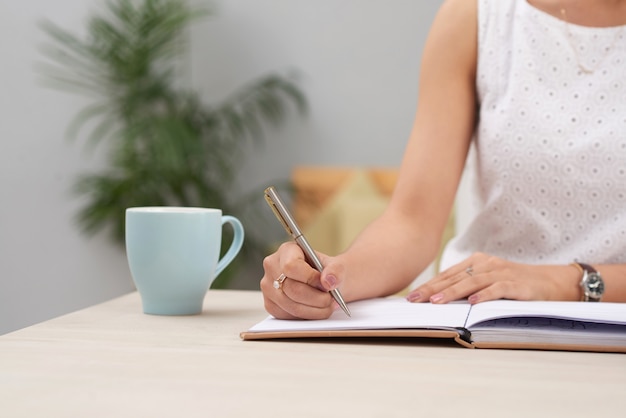 Unrecognizable woman in dress sitting indoors at desk and writing in journal