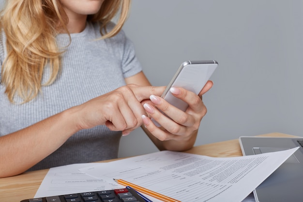 Foto gratuita la donna irriconoscibile tiene in mano il moderno telefono cellulare, si siede al tavolo di lavoro, circondata da documenti, laptop, matita, calcolatrice, scarica le informazioni necessarie da internet. lavoro e tecnologia