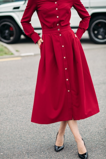 Unrecognizable woman in red dress with golden buttons and black heels posing in the street with hands in pockets of her skirt.