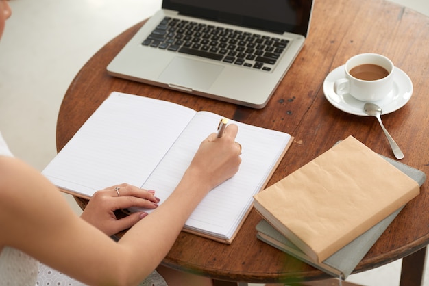 Unrecognizable woman sitting in cafe with laptop and writing in journal