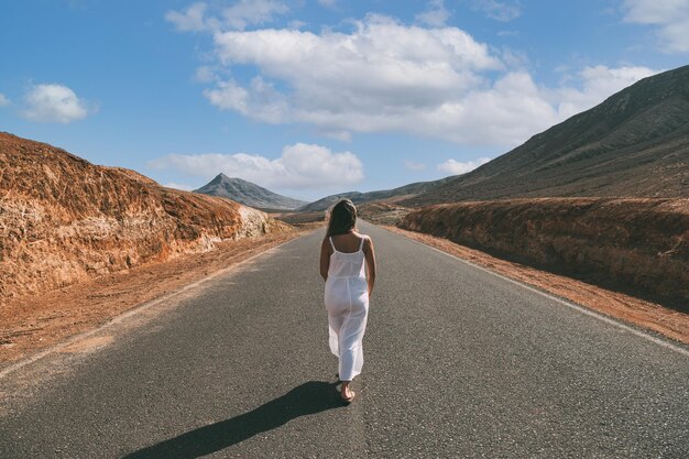 Unrecognizable woman walking on road near hills under cloudy sky