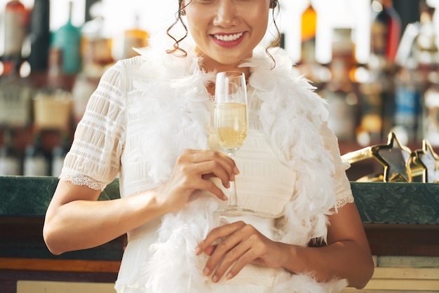 Unrecognizable woman with white feathered boa holding glass of champagne in bar