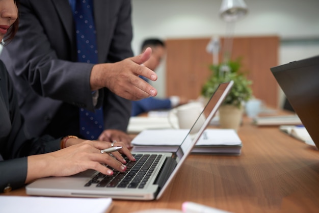 Unrecognizable woman working on laptop in office and man standing behind, watching and pointing