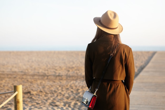 놀라운보기를 고민하는 세련된 모자, 코트 및 어깨 가방을 입고 인식 할 수없는 젊은 여성