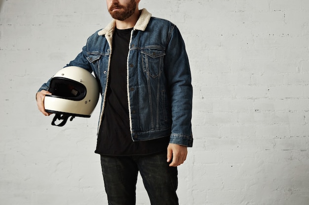 Free photo unrecognizable young motor biker wears shearling denim jacket and black blank henley shirt, hugs his vintage beige motorcycle helmet, isolated in center of white brick wall