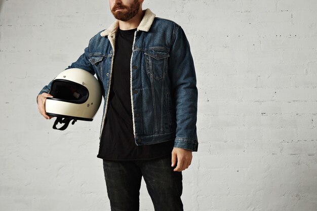 Unrecognizable young motor biker wears shearling denim jacket and black blank henley shirt, hugs his vintage beige motorcycle helmet, isolated in center of white brick wall