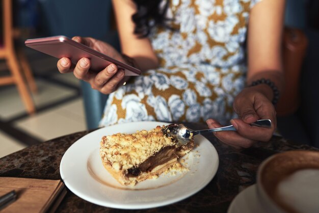 Unrecognizable young woman sitting in cafe, using smartphone and eating crumble pie