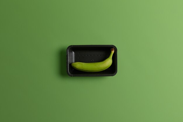 Unripe green banana on black tray. Tropical fruit for your consumption. View from above. Healthy lifestyle and nutrition. Fruit and food concept. Single banana collected from orchard. Vivid background