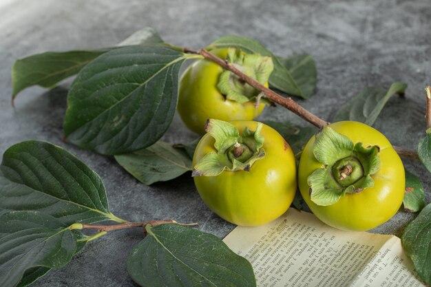 Unripe persimmons with leaves and book page on marble surface