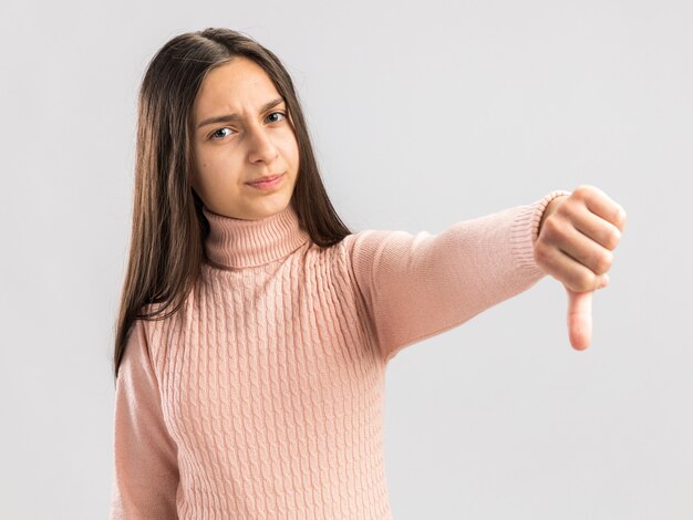 Unsatisfied pretty teenage girl looking at front keeping thumb down isolated on white wall