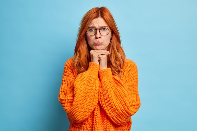 Unsatisfied sad woman with red hair keeps hands under chin feels unhappy purses lower lips looks directly dressed in knitted jumper.