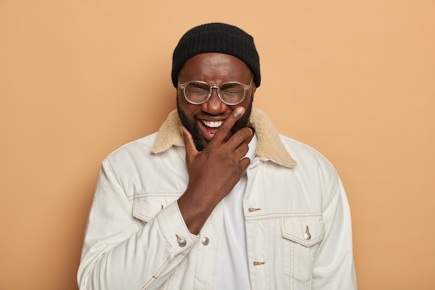 Free photo unshaven adult man with dark skin, laughs happily, holds chin, stands carefree