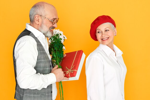 Unshaven bald elderly man wearing elegant clothes holding bunch of daisies and bof of chocolate, making birthday present to his charming wife