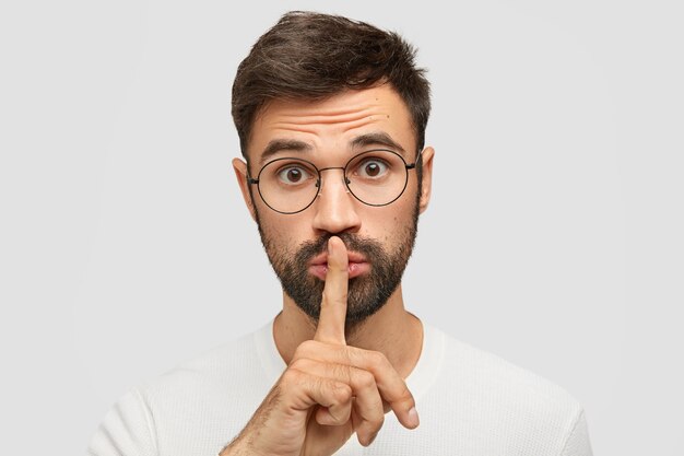 Unshaven Caucasian male makes silence gesture, asks to be quiet as someone sleeps, wears spectacles, has trendy haircut, isolated over white wall. People, conspiracy and secrecy concept