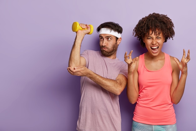 Unshaven guy raises arm with dumbbell, does exercises for training muscles and irritated curly haired woman gestures angrily, dissatisfied by something