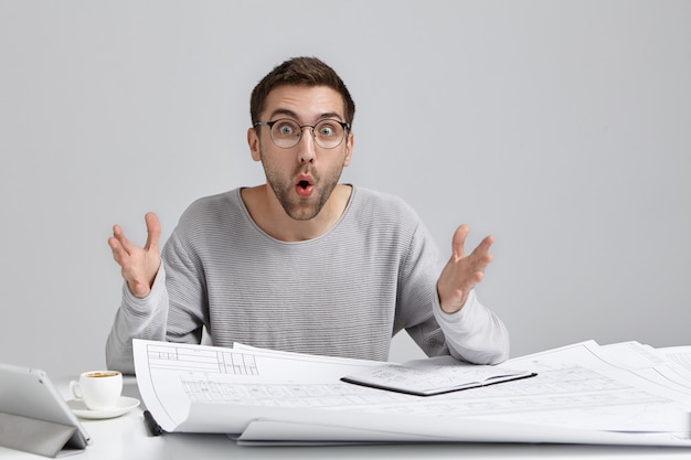 Free photo unshaven surprised male designer at work desk, gestures in shocked expression, looks bugged eyes