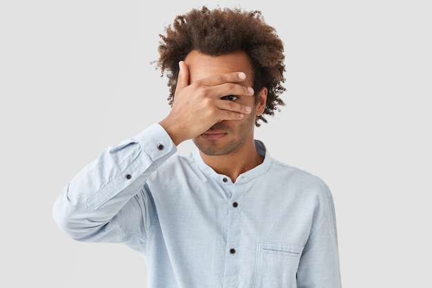 Unshaven young curly man covers eyes with palm, tries to be hidden, looks through fingers