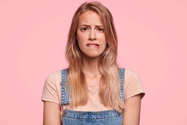 Unsure beautiful woman with displeased expression, bites lips, feels worried as has problems, wears casual t shirt with dungarees