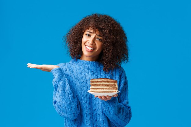 Unsure and clueless attractive african-american female in sweater, shrugging look uncertain, dont know eat or no, holding plate with tasty cake full calories, standing blue wall.