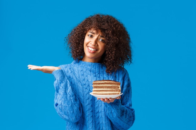 확실하지 않고 단서도없는 매력적인 아프리카 계 미국인 여성이 스웨터를 입고, 어깨를 으쓱하는 모습은 불확실하고, 먹거나 안하는지 몰라, 맛있는 케이크 전체 칼로리와 함께 접시를 들고, 파란색 벽에 서 있습니다.