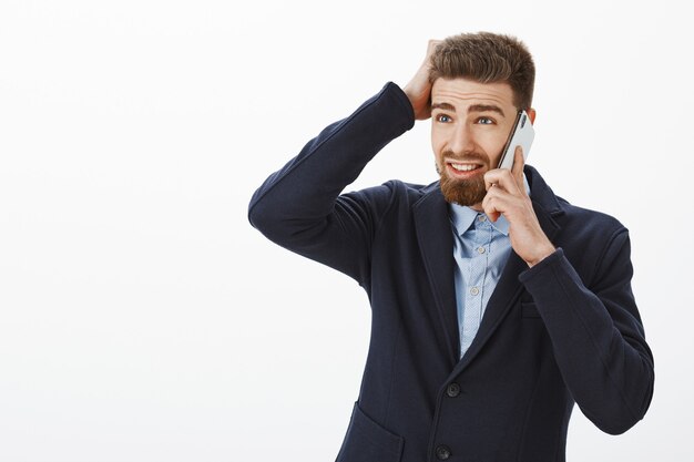 Unsure confused charming male entrepreneur in elegant suit with beard looking left with troubled face talking on smartphone scratching head apologizing via cellphone for working late over grey wall