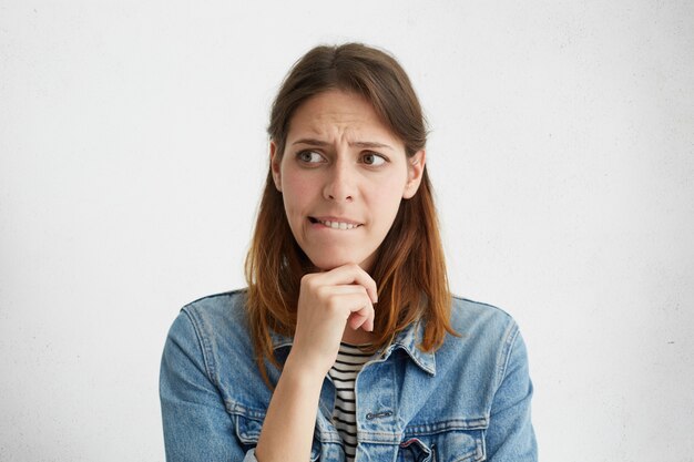 Unsure cute woman in casual clothes holding hand under chin biting lip looking aside while deciding what to do.