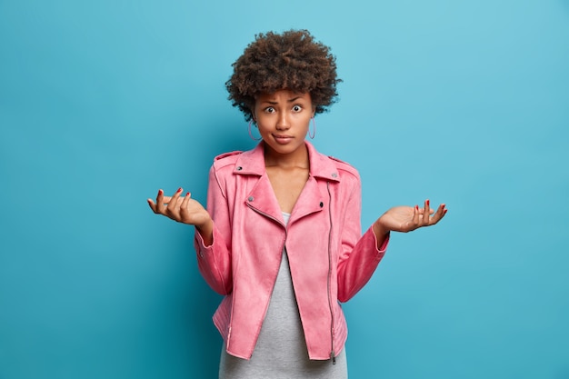 Unsure doubtful Afro American woman with curly hair spreads palms sideways, bothered by making choice, dressed in fashionable pink jacket, stands careless, cannot answer question, poses indoor
