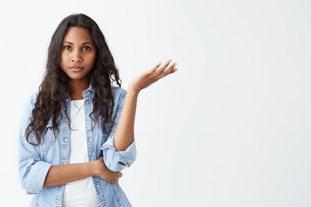 Unsure doubtful dark-skinned woman with black long wavy hair shrugging her shoulder in questioning gesture of uncertainty, having confused and dissatisfied look. Who cares