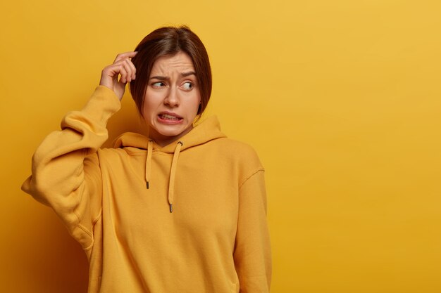 Unsure doubtful woman scratches head, has amnesia, makes tough decision, looks troubled aside, stands puzzled, clenches teeth, dressed in hoodie, poses against yellow wall, empty space for text