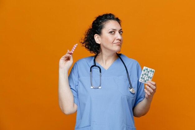 Неуверенная женщина-врач средних лет в униформе и со стетоскопом на шее показывает упаковки таблеток, глядя в камеру на оранжевом фоне
