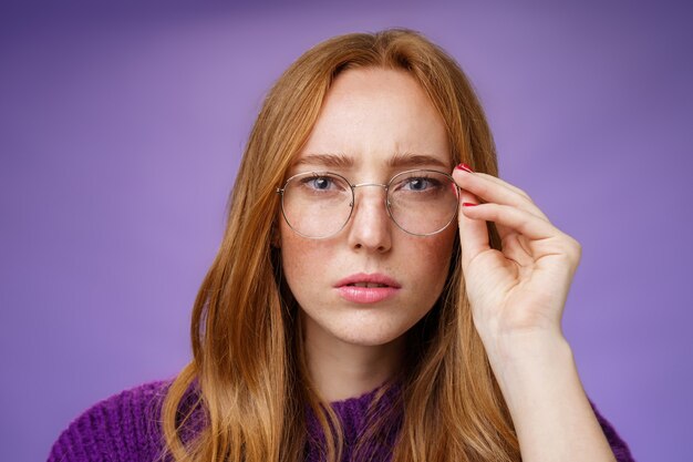 Unsure and serious-looking focused redhead woman frowning and squinting as looking in prescribed glasses at camera, touching frame reading sign, solving puzzle over purple background.