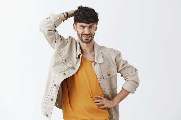 Unsure serious-looking stylish bearded guy posing against the white wall