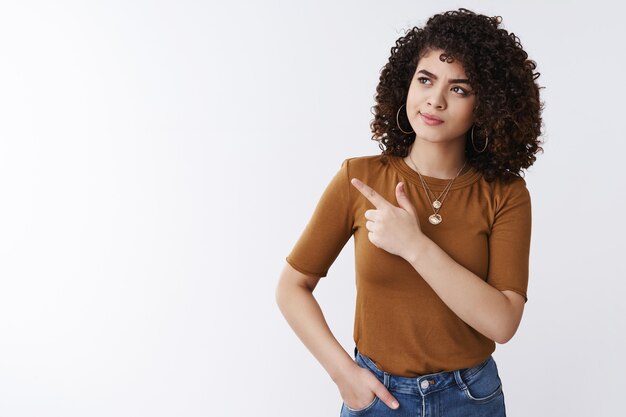 Unsure smart attractive curly-haired girl frowning smirking pointing left look thoughtful pondering important decision making choice standing perplexed unsure cannot remember white background