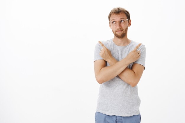 Unsure smart, creative male customer in glasses pursing lips and frowning looking away, crossing hands on chest pointing left and right making decision with doubts