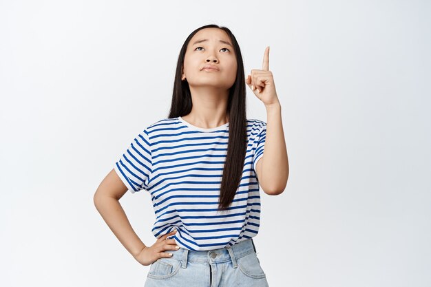 Unsure young asian woman pointing and looking up with troubled on white