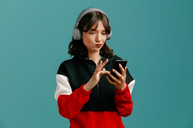파란색 배경에 격리된 휴대전화를 사용하여 헤드폰을 끼고 있는 불확실한 젊은 백인 여성