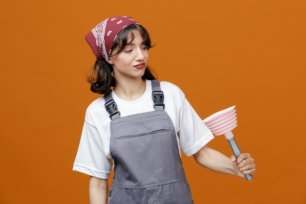 Неуверенная молодая женщина-уборщица в униформе и бандане держит и смотрит на поршень, изолированный на оранжевом фоне