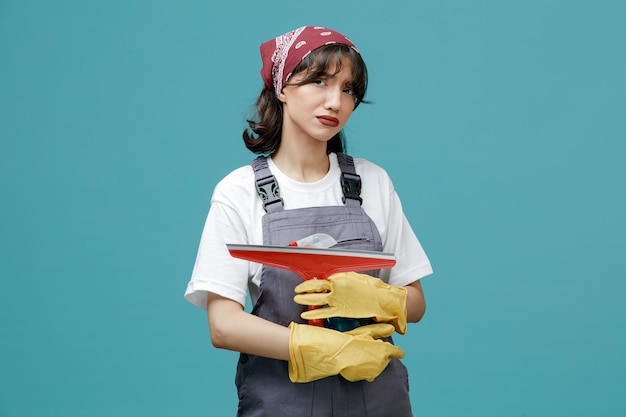 Unsure young female cleaner wearing uniform bandana and rubber gloves holding and hugging wiper and cleanser looking at camera isolated on blue background