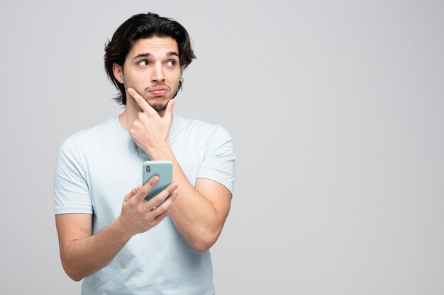 unsure young handsome man holding mobile phone keeping hand on chin looking at side isolated on white background with copy space