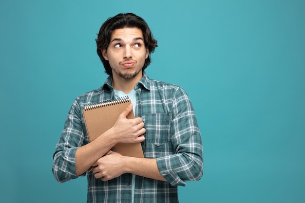 unsure young handsome man hugging note pad looking at side isolated on blue background with copy space