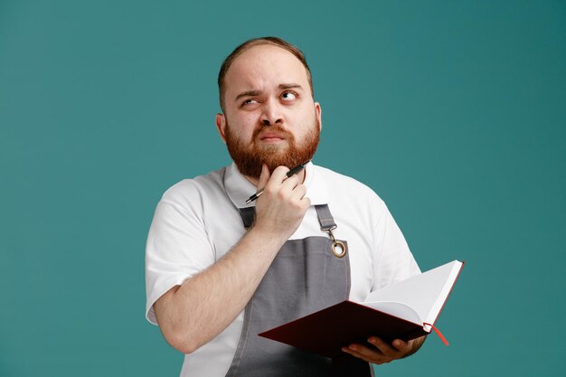 Unsure young male barber wearing white shirt and barber apron holding note pad and pen keeping hand on chin looking at side isolated on blue background