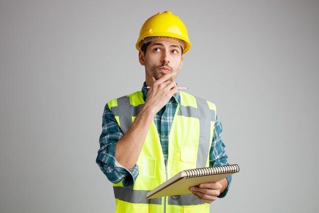 Неуверенный молодой мужчина-инженер в защитном шлеме и униформе держит блокнот и карандаш, держа руку на подбородке, глядя вверх на белом фоне