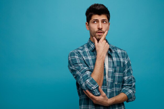 Unsure young man keeping hands on chin and under elbow looking at side isolated on blue background with copy space