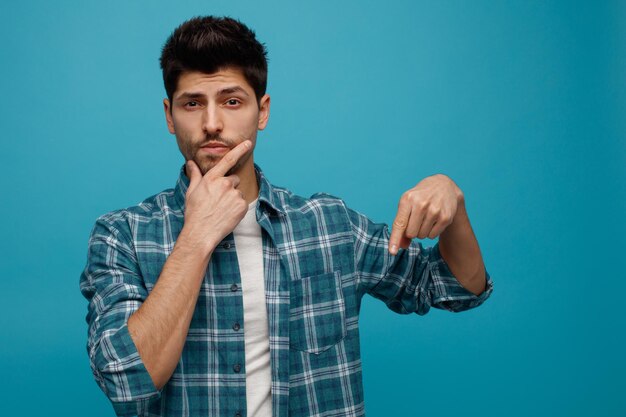 Unsure young man looking at camera keeping hand on chin pointing down isolated on blue background