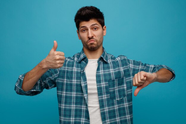 Unsure young man looking at camera showing thumbs up and down isolated on blue background