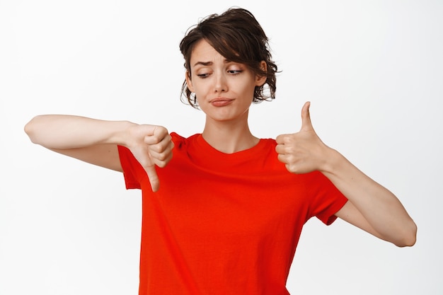 Unsure young woman making choice, show thumbs up and thumbs down, being indecisive, say no or yes, judging smth, standing on white.