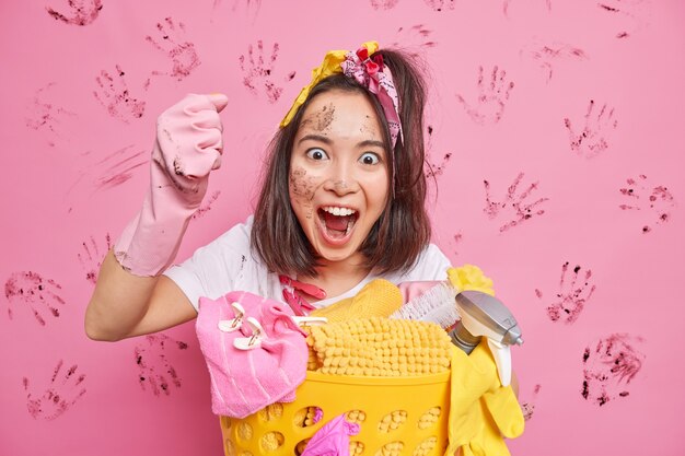 Неопрятная домохозяйка громко восклицает, сжимает кулак, делает уборку дома позы возле корзины с бельем с грязным лицом, изолированным над розовой стеной